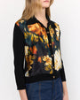 A person wearing a black cardigan with vibrant yellow, orange, and white floral print on the front, paired with blue jeans. The cardigan has solid black sleeves and collar, and buttons down the front. The background is plain.