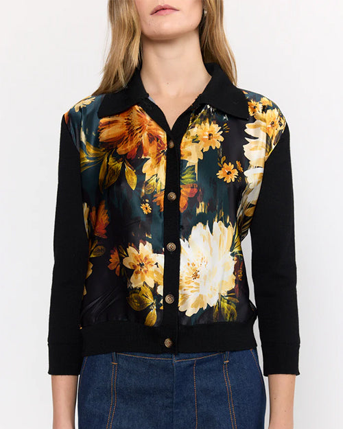 Front view of a person wearing a black cardigan with vibrant yellow, orange, and white floral print on the front, paired with blue jeans. The cardigan has solid black sleeves and collar, and buttons down the front. The background is plain.
