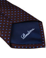 A back view of a necktie with a brown base color, with a light blue pattern dots. The tie features a navy blue interior and small white lettering. 