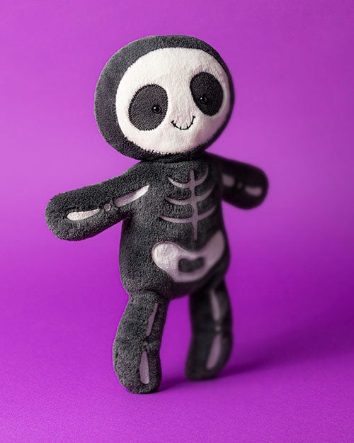 Plush toy designed as a cute skeleton with a black body and white bone details, including skull, ribcage, and pelvis, featuring large black eyes and a small smile, set against a purple background.