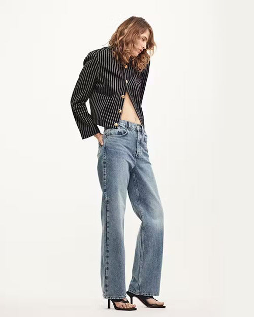 A person stands wearing light-wash, high-waisted, straight-leg jeans with a relaxed fit. The jeans feature a button fly closure and classic five-pocket design. The person is also wearing a black and white striped shirt with an open midriff and black high-heeled shoes. The background is plain and white, emphasizing the casual yet stylish outfit centered around the denim jeans.