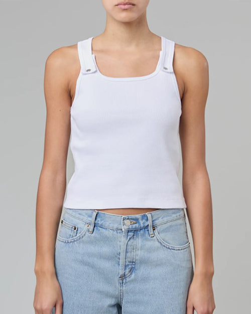 A model is wearing a white tank top with button snap straps that close at the upper chest. The model pairs the shirt with light blue jeans.