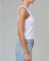 A side view of a model wearing a white tank top with button snap straps that close at the upper chest. The model pairs the shirt with light blue jeans.