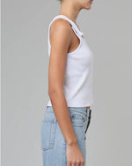 A side view of a model wearing a white tank top with button snap straps that close at the upper chest. The model pairs the shirt with light blue jeans.