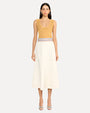A dark-haired model wearing a sleeveless mustard yellow top with a scoop neckline, tucked into a white midi skirt with an embroidered blue and red waistline. The model is wearing silver open-toed heels to match and is standing in front of a white background.