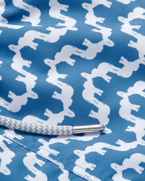 A close-up of swim shorts with a repeating pattern of white silhouettes. There is a white drawstring with a metal aglet at its end.