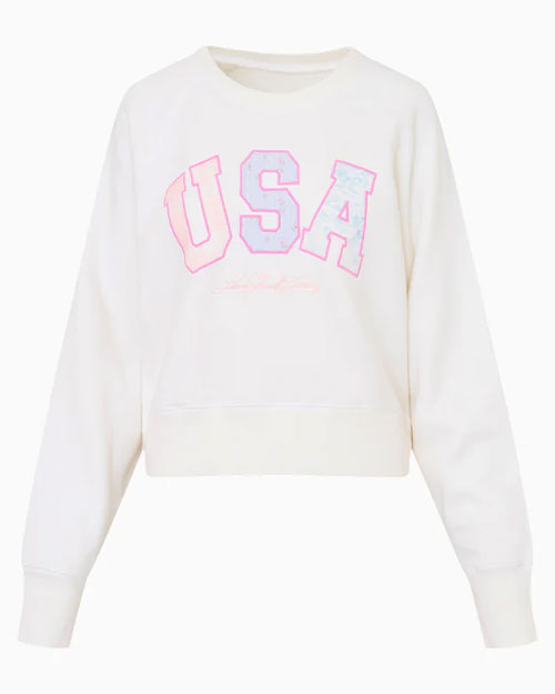 A white sweatshirt with the text "USA" printed on the front in large, bold letters. The letters are outlined in pink and filled with a light blue, light orange, and silver pattern. Below the "USA" text, there is smaller cursive text.
