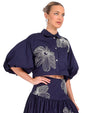 "A person wearing a navy blue two-piece outfit with a cropped button-up shirt featuring puffy sleeves and a high-waisted skirt. Both pieces are adorned with large white floral embroidery.