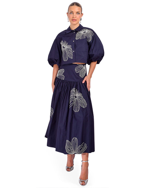 A person wearing a navy blue two-piece outfit with a cropped button-up shirt featuring puffed sleeves and a high-waisted, full-length skirt. The outfit is adorned with large white floral patterns, and the person is wearing silver high-heeled sandals.