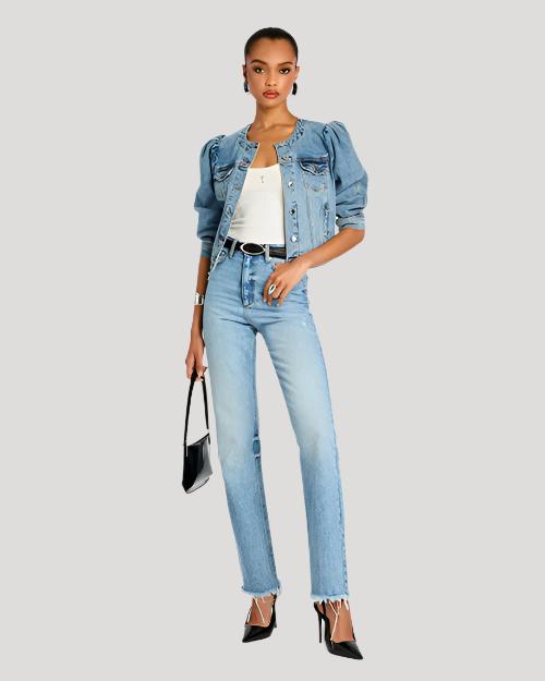 A person stands against a plain background, wearing a denim jacket with puffed sleeves, high-waisted jeans, and black pointed-toe heels. The individual is accessorizing with a thin belt and carries a black shoulder bag.