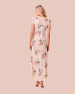 Back view of a person standing against a light beige background, wearing a floral dress with short sleeves and a V-neckline that reaches down to the ankles. The dress is primarily white with pink and blue floral patterns throughout.