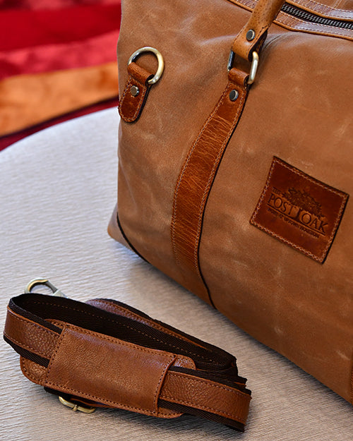 A close-up image of a brown leather bag with a detachable strap placed on a light-colored surface. The bag has metal rings and buckles, and a leather patch with the text 'POST OAK' embossed on it.