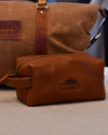 A travel kit with a The Post Oak Hotel logo embossed on the side on a beige surface. In the background, there is a brown duffle bag.