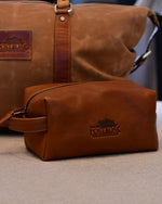 A travel kit with a The Post Oak Hotel logo embossed on the side on a beige surface. In the background, there is a brown duffle bag.