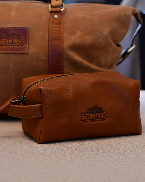 A travel kit with a The Post Oak Hotel logo embossed on the side on a beige surface. In the background, there is a brown duffle bag.
