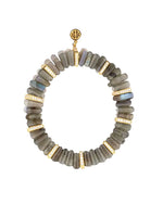 Bracelet made of cylindrical labradorite beads in grayish-green tones with iridescent flashes, accented by gold spacers encrusted with small white gemstones. A round gold charm with an intricate design is positioned at the top.