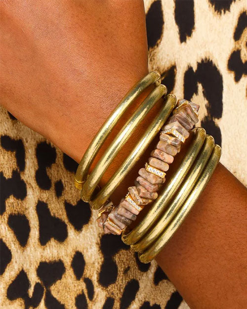 Close-up of a wrist wearing multiple bracelets, including four gold bangles and one made of irregularly shaped beige stones with small gold accents. The background displays a leopard print pattern.
