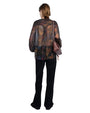 Person standing with their back to the camera, wearing a sheer multicolored long-sleeved blouse, black pants, and carrying a brown shoulder bag on the left side.