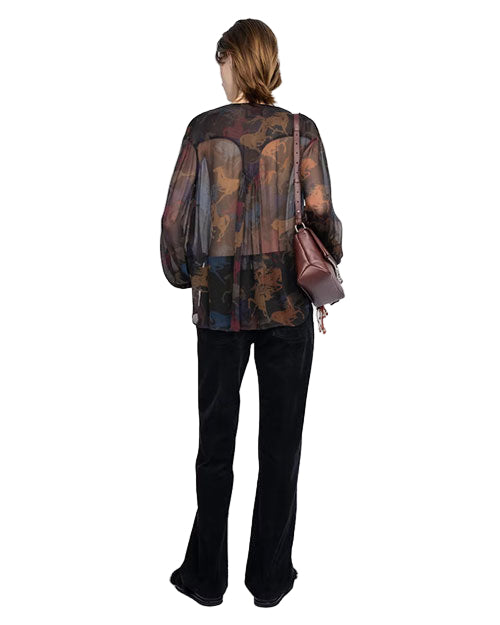 Person standing with their back to the camera, wearing a sheer multicolored long-sleeved blouse, black pants, and carrying a brown shoulder bag on the left side.