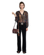 Person wearing a sheer long-sleeved blouse with a lace-up neckline, black pants, and black sandals, holding a small object in the right hand and carrying a maroon handbag with decorative elements over the left shoulder. 
