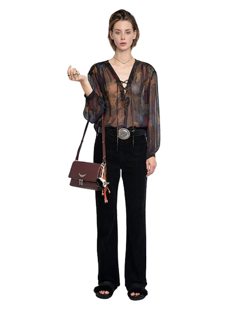 Person wearing a sheer long-sleeved blouse with a lace-up neckline, black pants, and black sandals, holding a small object in the right hand and carrying a maroon handbag with decorative elements over the left shoulder. 