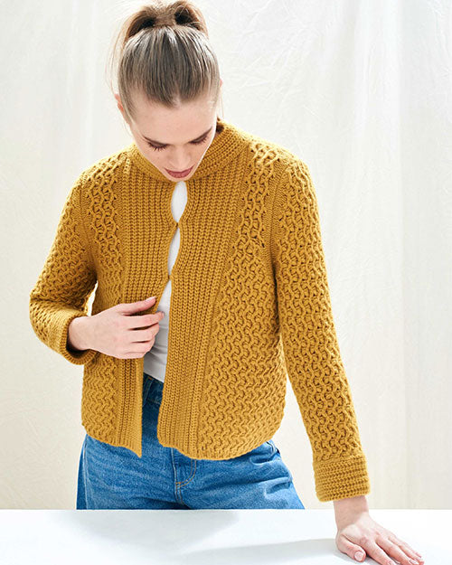 Person wearing a mustard yellow knitted cardigan with intricate patterns, long sleeves, and an open front revealing a white top, paired with blue jeans. Her hair is tied in a ponytail and she is facing down. 
