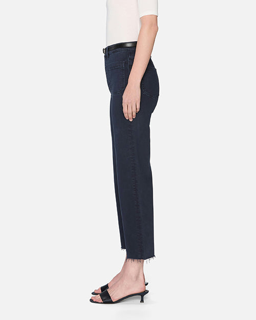 The side view of a person wearing a white short-sleeve top tucked into high-waisted, navy denim jeans with a black belt. The jeans have zipper and button closure, two large front pockets, and frayed hems at the bottom. The person is also wearing black open-toe sandals.