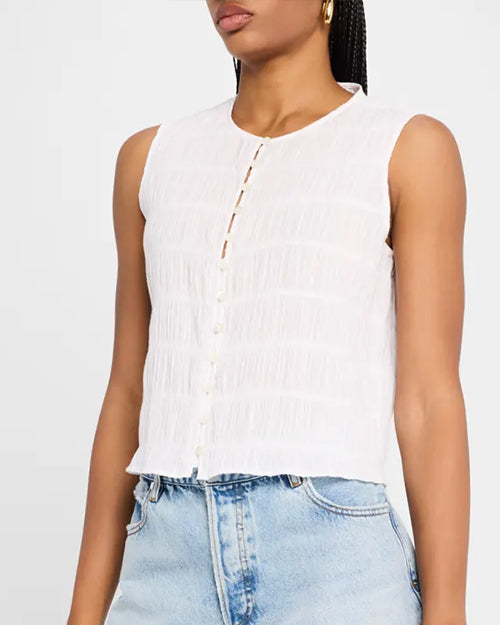 A model wearing a sleeveless white blouse with a textured, horizontal stripe pattern, a button-down front with small white buttons and a collarless neckline. The model is also wearing light blue jeans and is standing against a white background.