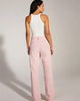 The backside view of a brunette model wearing a white sleeveless top and light pink jeans, standing against a grey background.