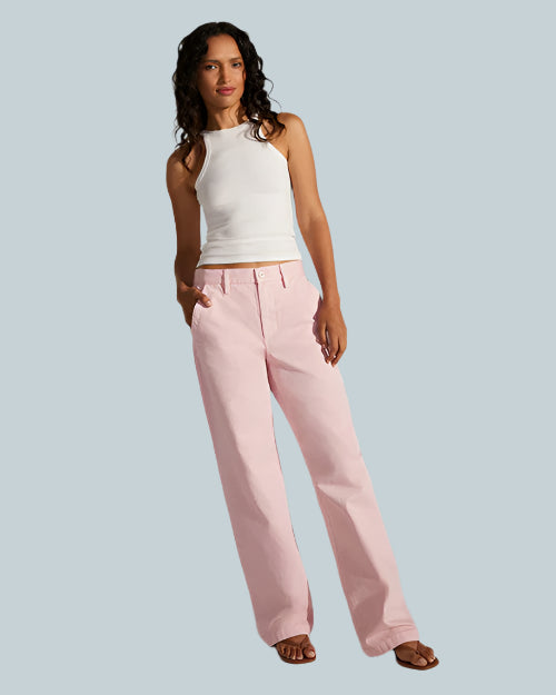 A brunette model with her hand in her pocket, is wearing a white sleeveless top with light pink jeans and brown open-toed sandals. The model is standing against a grey background.