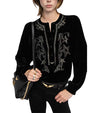 Person wearing a black velvet blouse with star and floral embroidery, buttoned front, and black pants, carrying a black handbag with gold chain details and wearing multiple rings.