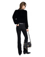 Back view of a person wearing a black long-sleeve top, black pants, and black shoes, holding a black handbag with gold accents in the left hand. 