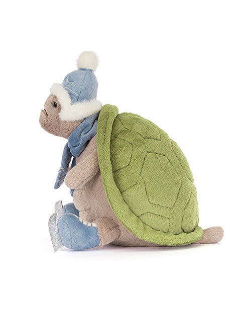Plush turtle toy with a green hexagonal-patterned shell and light brown body, wearing a blue winter hat with white pom-pom and fur trim, a matching blue scarf, and blue ice skates.