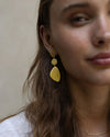 A close-up of a person with long, wavy brown hair wearing a white top, a gold necklace, and gold earrings on the left ear. 