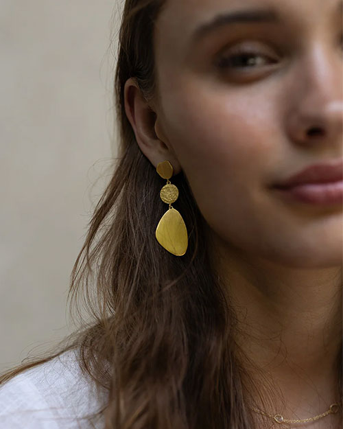 A close-up of a person with long, wavy brown hair wearing a white top, a gold necklace, and gold earrings on the left ear. 