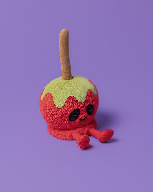 Plush toy shaped like a candy apple with a red body, green dripping candy detail on top, a stick protruding from the top, large black eyes, and small red feet, set against a solid purple background.