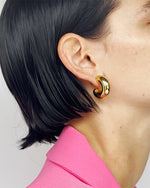 Close-up of a person’s left side profile with golden earring with a unique spiral design that resembles a c. The earring is worn on the earlobe against the backdrop of short black hair and a vibrant pink garment.
