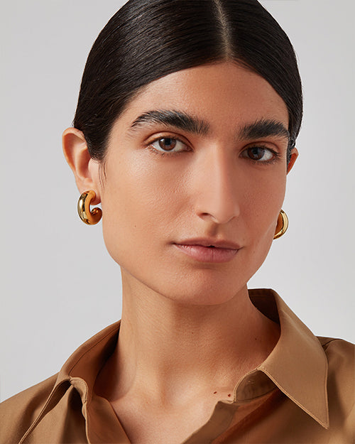 Close-up of gold hoop earrings with, medium-sized and snugly fitting around the earlobes. The person wearing them has a light brown blouse with the collar slightly turned up.