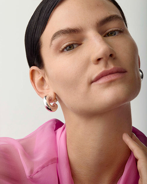 Close-up of model from the shoulders up. They are wearing a pink shirt, and their left earlobe showcases an earring that is medium sized, chunky, silver hoop.