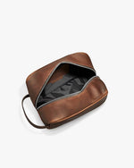 Open brown leather dopp kit with a zipper. The interior is black with visible stitching and compartments, and there’s a side handle on the right.