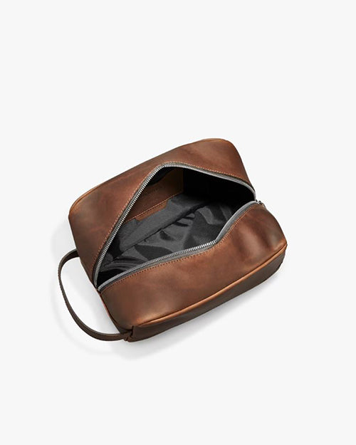An open, empty brown leather dopp kit with a zipper on a white background. The interior is black with visible stitching and compartments, and there’s a side handle on the right. The kit is suitable for storing toiletries or grooming supplies.