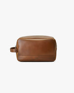 Brown leather dopp kit with a rectangular shape, a small handle on right side, and zipper on the top. The brand name is in small letters near the lower edge of the bag.