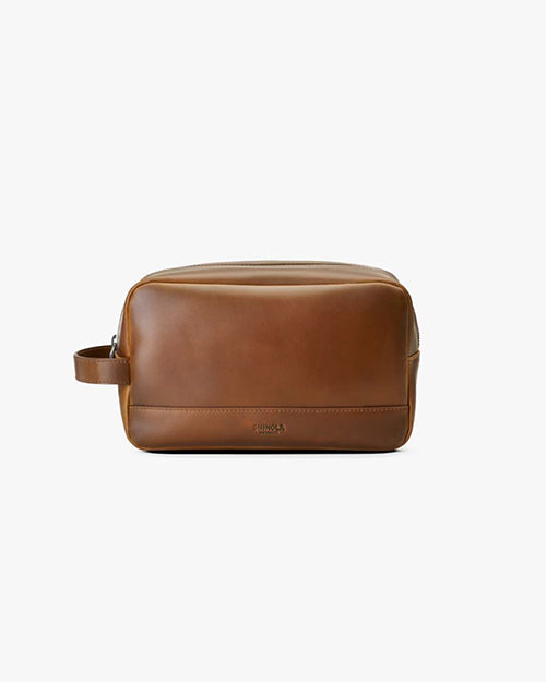 A single, brown leather dopp kit with a smooth texture and a visible handle on the right side. The kit has a rectangular shape with rounded edges and features a zipper closure on top. The brand name is embossed in small letters near the lower edge of the bag’s side facing the viewer. 