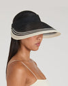 A person with long dark hair wearing a wide-brimmed visor hat with alternating black and white stripes. The hat covers the upper part of the person's face. They are dressed in a sleeveless, light-colored top, and the background is plain light gray.