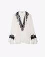 A full view of a white blouse with black lace detailing with scalloped edges around the V-neck neckline and around the lower end of the long sleeves with flared cuffs. The background is plain white.