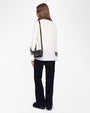 A full body backside view of a brunette model with her hair swept to the side, wearing a white blouse with black lace detailing around the lower end of the long sleeves. She is wearing black pants to match, black shoes, and a small black purse. The background is plain white.