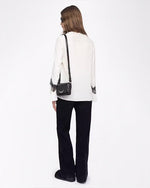A full body backside view of a brunette model with her hair swept to the side, wearing a white blouse with black lace detailing around the lower end of the long sleeves. She is wearing black pants to match, black shoes, and a small black purse. The background is plain white.
