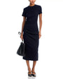 Person wearing a fitted, short-sleeved navy blue dress with ruched detailing at the waist, holding a black handbag and wearing black ankle-strap flats.