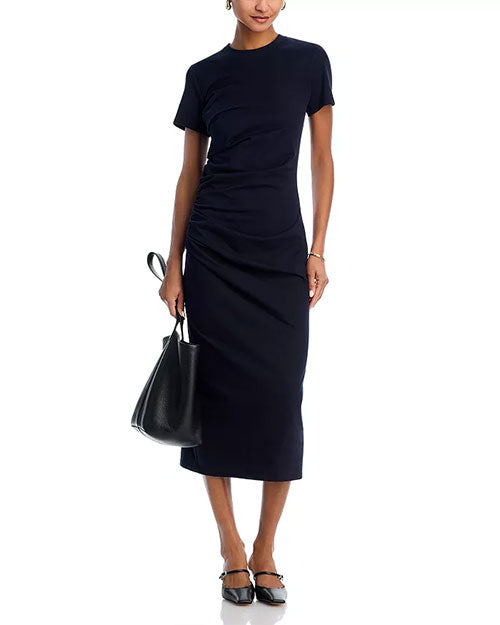Person wearing a fitted, short-sleeved navy blue dress with ruched detailing at the waist, holding a black handbag and wearing black ankle-strap flats.