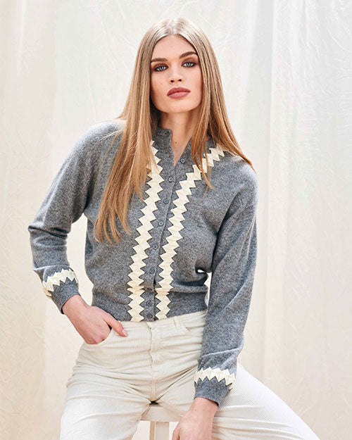Person sitting on a stool wearing a grey cardigan with white zigzag patterns on the front and cuffs, paired with light-colored pants. 